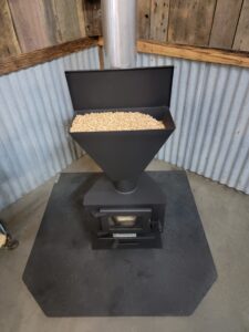 Gravity-fed pellet stove with hopper full of wood pellets installed on a hexagonal hearth pad in a corner of a metal-walled workshop.