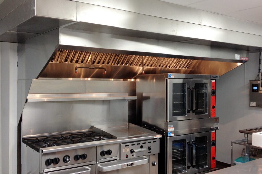 Stainless steel commercial kitchen exhaust hood with baffle filters above a gas range, griddle, and double convection ovens.