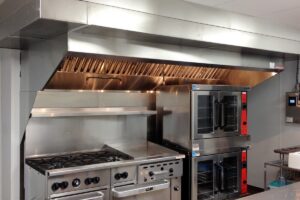 Stainless steel commercial kitchen exhaust hood with baffle filters above a gas range, griddle, and double convection ovens.