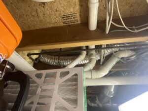 Dryer vent duct rerouted between floor joists in an unfinished basement, running past PVC piping and a furnace filter.